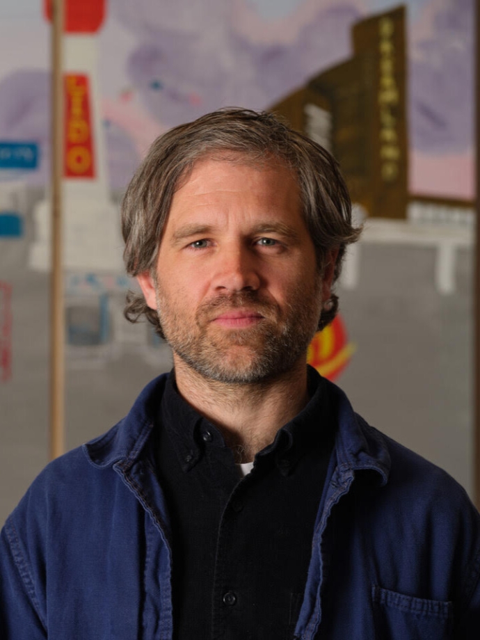 Man with wavy grayish-brown hair wearing a dark blue jacket and black shirt, standing in front of classroom.